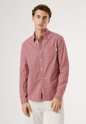 Camisa de cuadros rojos de manga larga, fabricada en algodón, con cuello abotonado y bolsillo, presenta botones blancos y una pequeña etiqueta en el costado.