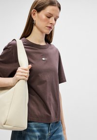 Brązowy bawełniany t-shirt z krótkim rękawem, z małym czarno-białym logo na piersi. Modelka trzyma kremową torebkę na ramię.