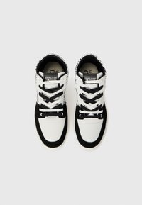 Ténis de cano alto brancos e pretos com atacadores pretos e etiqueta "Versace Jeans Couture" na lingueta, vistos de cima sobre um fundo simples.