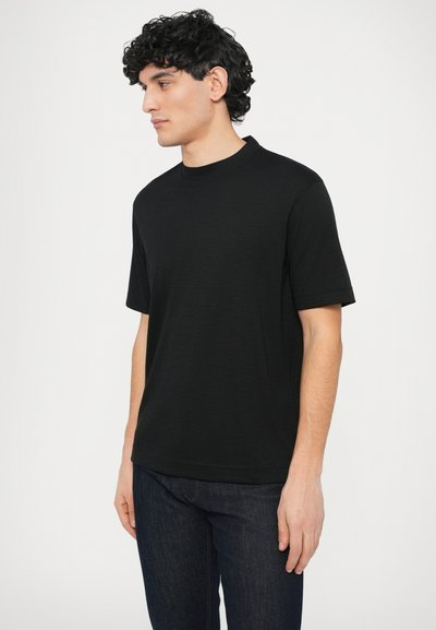 Fekete rövid ujjú póló kerek nyakkivágással, sima textúrával és visszafogott márkajeléssel; sötétkék farmernadrággal párosítva.