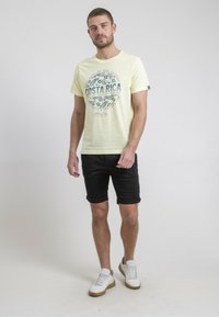 T-shirt jaune à manches courtes avec un graphisme "COSTA RICA", associé à un short noir roulé et des baskets blanches avec des accents bruns.