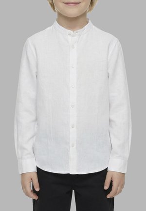 Bambino che indossa una camicia bianca a maniche lunghe con colletto alla mandarino e pantaloni neri su uno sfondo grigio uniforme.