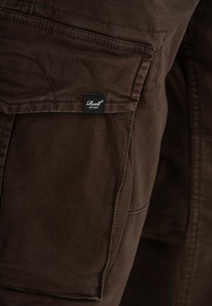 Brown fabric pants with a prominent side pocket and a black label reading "Reell est. 1997." Features sturdy stitching and a soft texture.