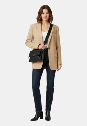 Donna con blazer beige, top bianco, jeans scuri, stivaletti neri e borsa a tracolla in pelle nera con tracolla larga.