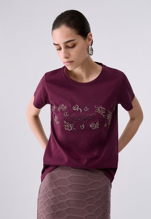 Donna con capelli scuri che indossa una maglietta viola ricamata con fiori e una gonna mauve strutturata, in piedi con le mani dietro la schiena su uno sfondo semplice.