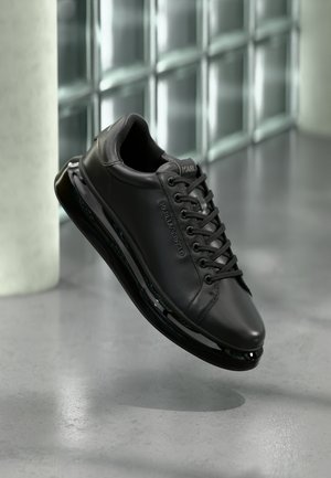 Schwarzer Karl Lagerfeld Ledersneaker mit schwarzen Schnürsenkeln, der über einem grauen Boden und einer Wand aus Glasbausteinen schwebt.
