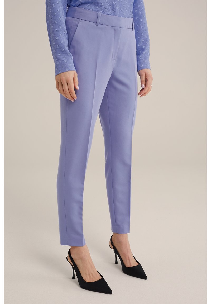 Femme portant un pantalon violet clair sur mesure longueur cheville et des talons hauts noirs pointus, debout devant un fond uni.