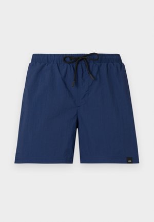 Homme debout, les mains dans les poches, portant une chemise noire à manches courtes boutonnée, un short bleu et des chaussures bateau en cuir marron.