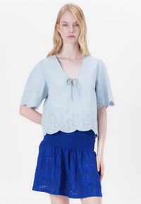 Ανοιχτό μπλε κοντομάνικο μπλουζάκι με V-λαιμό και περιθώριο με κύματα, με κεντητά σχέδια. Συνδυασμένο με μια υφή, φρατζόν σκοτεινής μπλε φούστας.