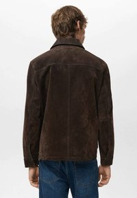 Smeđa jakna od brušene kože s ovratnikom, vidljivim šavovima i ravnim krojem. Ima bočne džepove i uparena je s plavim trapericama.