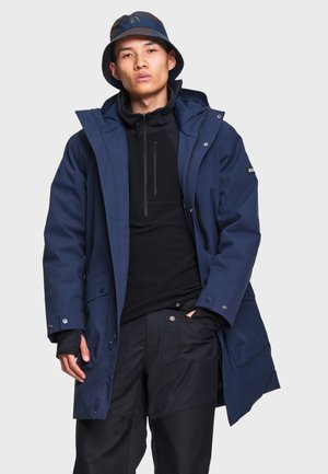 Jovem vestido com casaco azul-marinho com capuz, camisola preta com fecho, calças pretas e chapéu tipo bob azul, parado com uma mão no bolso.