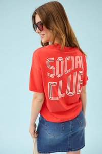 Femme portant de grandes lunettes de soleil noires, un t-shirt rouge avec le texte blanc "SOCIAL CLUB" dans le dos, et une jupe en jean bleue, sur un fond bleu clair.