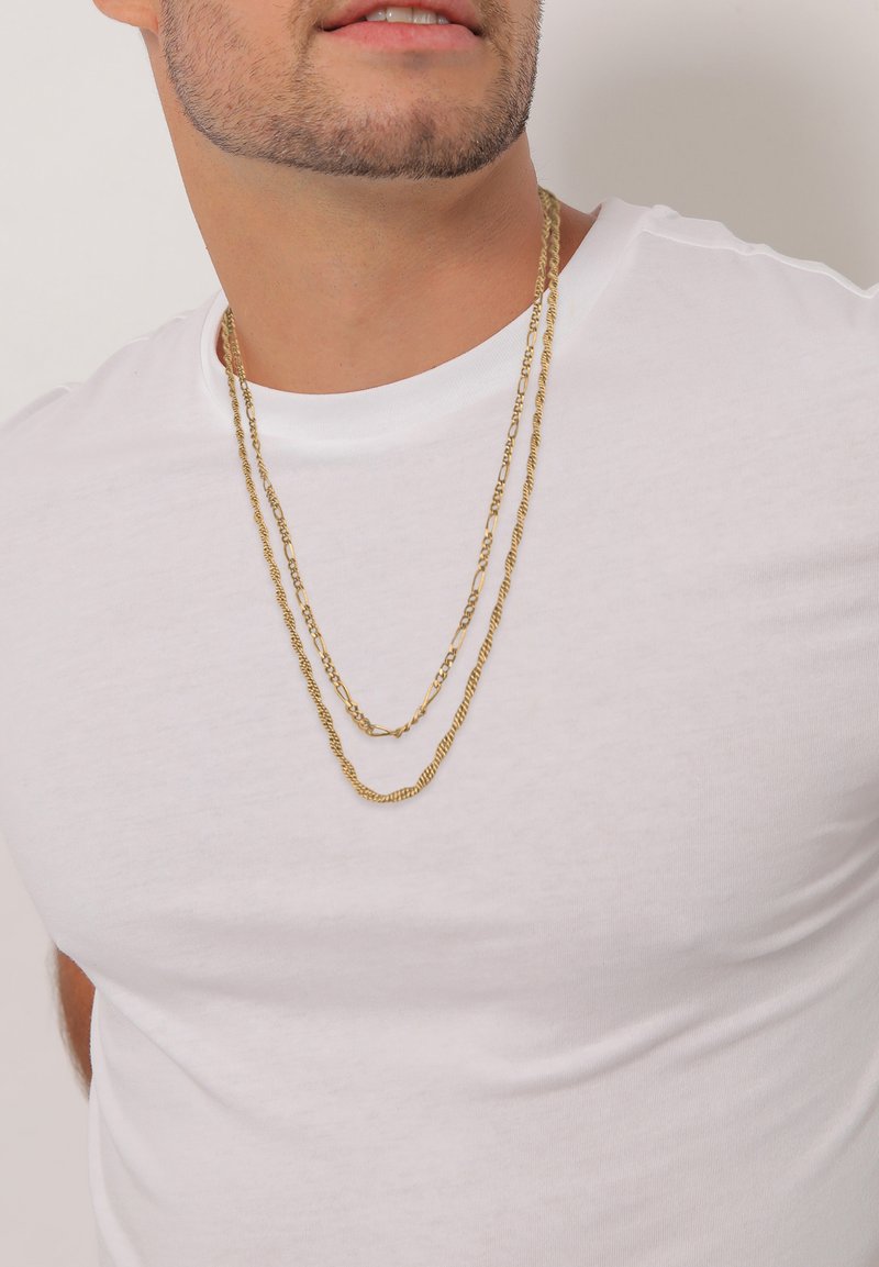 Gouden metalen kettingen in verschillende lengtes, gelegd over een wit T-shirt, met gedraaide en platte schakels.