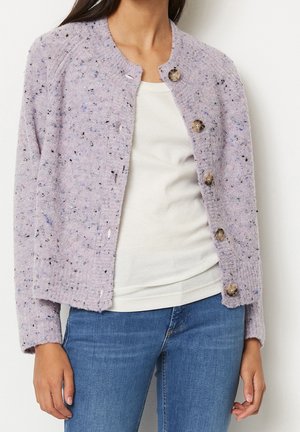 Femme portant un cardigan boutonné texturé lavande sur un haut blanc et un jean bleu, montrée du cou aux hanches.