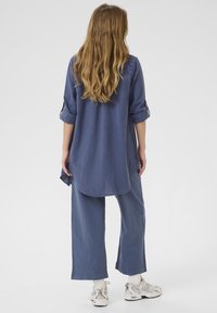 Mujer con cabello largo y ondulado, vistiendo una camisa azul de mangas arremangadas y pantalones azules sueltos, calcetines blancos y zapatillas blancas, de pie de espaldas.