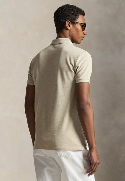 Man draagt een beige poloshirt met korte mouwen en witte broek, staat zijwaarts met een zonnebril tegen een eenvoudige neutrale achtergrond.