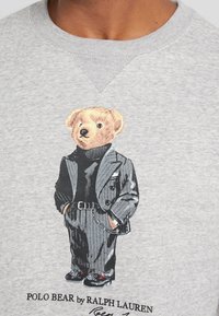 Grå sweatshirt med en broderad björn i en randig kostym, med hög hals och sneakers, texten nedan lyder "POLO BEAR av RALPH LAUREN."