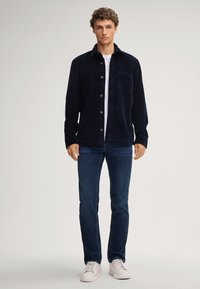Giacca camicia in corduroy navy con chiusura a bottoni e tasca, abbinata a jeans blu e sneakers bianche. Texture liscia, vestibilità classica.