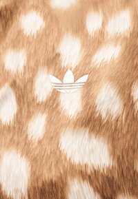 Tissu marron avec des taches blanches irrégulières ressemblant à de la fourrure d’animal, avec un petit logo Adidas trefoil blanc brodé au centre.