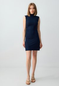 Marineblauwe mouwloze jurk met een hoge hals en een gestructureerd patroon, aansluitende silhouet en een knielange snit. Gedragen met simpele sandaaltjes.