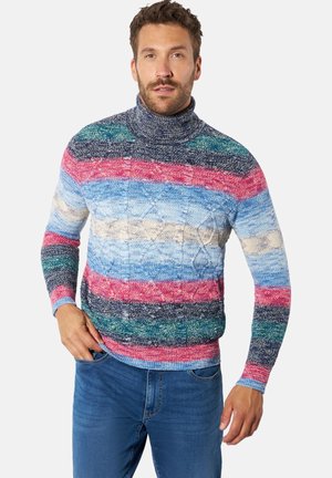 Mann trägt einen mehrfarbig gestreiften Rollkragenpullover mit blauer Jeans, steht mit einer Hand in der Tasche vor weißem Hintergrund.
