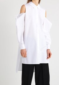 White long-sleeve shirt dress with cold shoulder cutouts and extended hem, worn with loose black pants on a standing person.