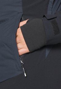 Veste noire et bleu foncé avec une poche zippée. Dotée d'un poignet texturé et d'un tissu lisse, conçue pour la fonctionnalité et le confort.