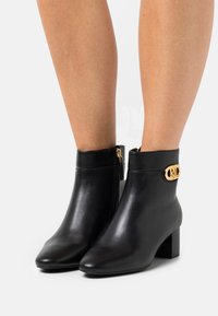 Botas negras de cuero hasta el tobillo con punta afilada, acentos de hardware dorado y tacón cuadrado. Textura suave, cierre de cremallera en la parte posterior.