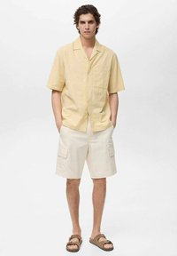 Kortärmige, gele leinen shirt met een zak, gecombineerd met beige cargoshorts met zijzakken, afgemaakt met bruin sandaal.