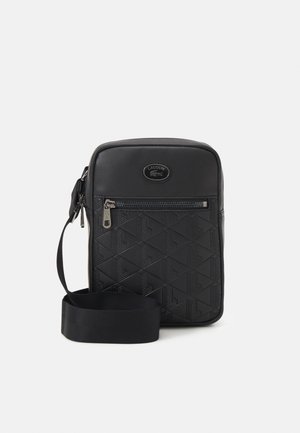 Sac bandoulière noir Lacoste avec motif géométrique embossé, poche avant zippée et bandoulière réglable sur fond blanc.