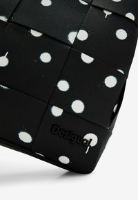 Czarna pleciona torba z białymi kropkami, o teksturowanej powierzchni i z czarnym logo marki. Kompaktowy i nowoczesny design, odpowiednia do różnych zastosowań.