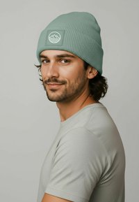 Mint green ribbed beanie with a square patch featuring a wave design. The model wears a grey short-sleeve shirt in a neutral setting.