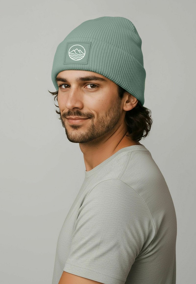 Mint green ribbed beanie with a square patch featuring a wave design. The model wears a grey short-sleeve shirt in a neutral setting.