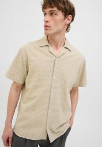 Camicetta beige a maniche corte con motivo geometrico bianco, chiusura a bottoni sul davanti e colletto. Realizzata in tessuto leggero, presenta una vestibilità comoda.