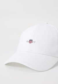 Witte baseballpet van katoen met een gebogen rand. Voorzien van een geborduurd logo met rode en blauwe accenten aan de voorkant. Glad van textuur.