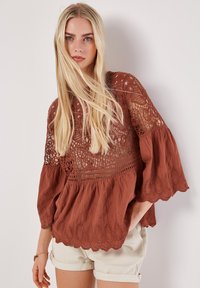 Blouse couleur rouille avec détails en dentelle, manches évasées et ourlet festonné. Fabriquée à partir d'un tissu texturé avec des motifs complexes.