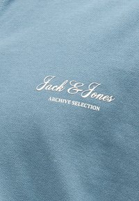 Ανοιχτό μπλε ύφασμα με λευκό κεντητό κείμενο που γράφει «Jack & Jones ARCHIVE SELECTION» σε υφή επιφάνειας.