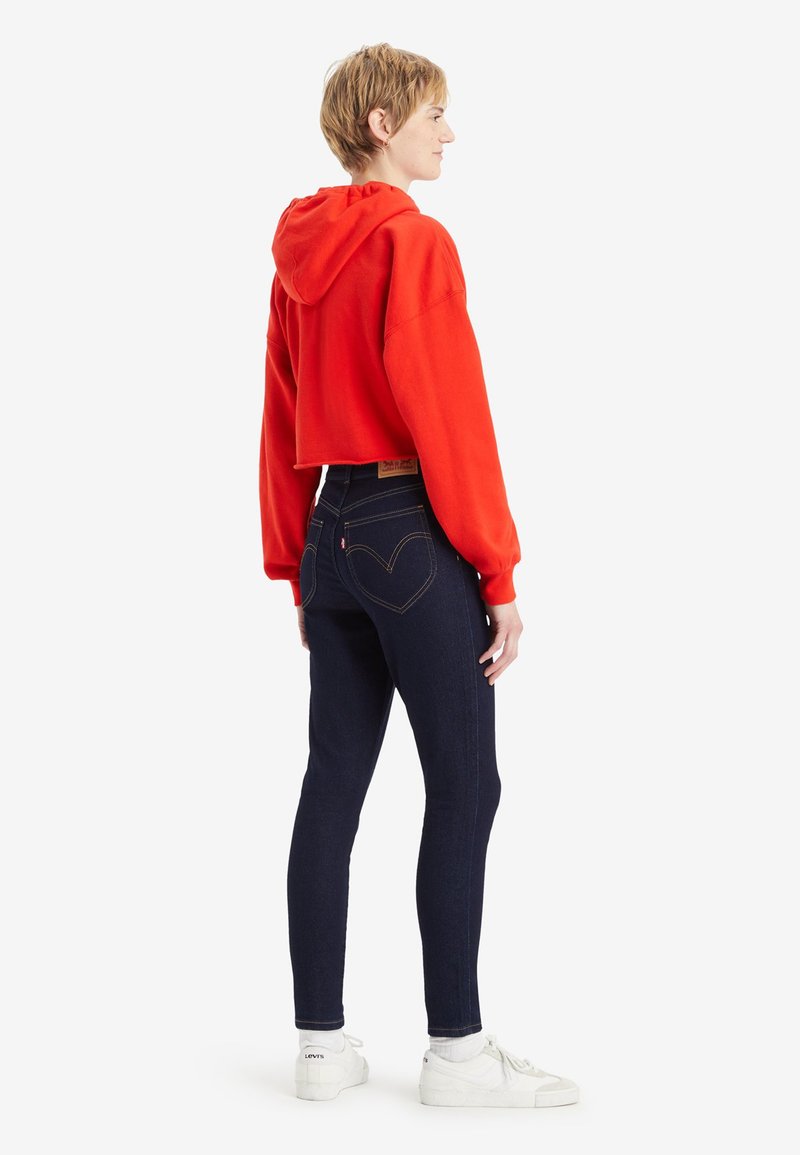 Haut à capuche rouge coupé avec des manches longues, accompagné de jeans taille haute bleu foncé avec un design de poche arrière en forme de cœur et des baskets blanches.