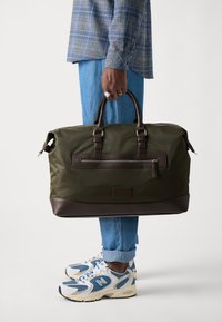 Olive green nylon duffel bag with brown leather accents, two top handles, and a front zipper pocket, held by a person in denim.