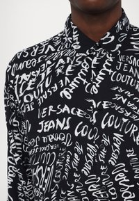 Schwarzes Hemd mit einem auffälligen, weißen Graffiti-Stil Druck der Wörter "Versace", "Jeans" und "Couture". Mit Kragen und Knopfverschluss.