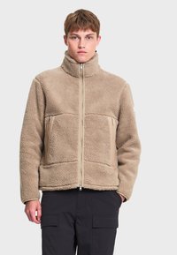 Jeune homme aux cheveux courts portant une veste polaire beige avec un col montant et des poches avant zippées, associée à un pantalon noir.