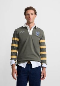 Camiseta de rugby de manga larga verde con cuello blanco, rayas amarillas en las mangas y logo de escudo bordado en el pecho. Corte clásico y tejido texturizado.