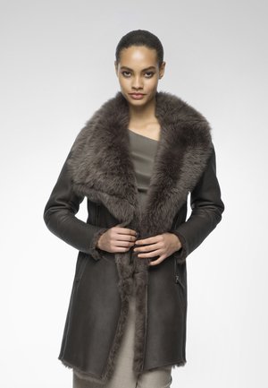 Manteau en cuir marron avec un large col en fourrure, présentant une texture lisse, des poches zippées et une coupe ajustée. Le manteau est longueur en dessous des hanches.