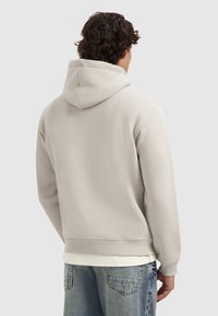 Mann mit lockigem Haar, der einen hellgrauen Hoodie und blaue Jeans trägt, von hinten gegen einen schlichten hellen Hintergrund gezeigt.