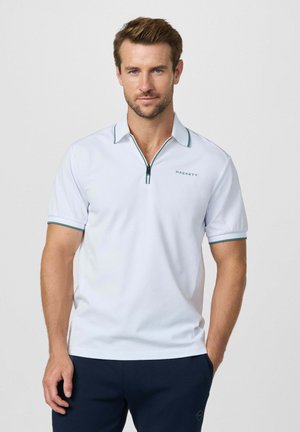 Hombre vistiendo una camisa polo blanca de manga corta con ribete verde y logo de "Hackett", combinada con pantalones azul marino, de pie frente a un fondo blanco.