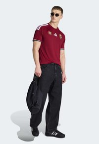 Vinrød atletik t-shirt med hvide striber, Adidas-logo og Ungarns våbenskjold. Matchende med sorte løstsiddende jeans og sort taske.