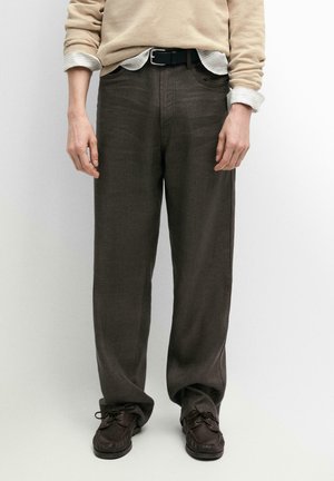 Uomo che indossa pantaloni marroni a taglio largo, cintura nera, maglione beige sopra una camicia a righe e scarpe stringate marrone scuro su uno sfondo bianco uniforme.