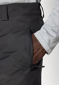 Main à la peau foncée dans une chemise à manches longues gris clair insérée dans la poche zippée latérale de pantalons noirs avec passants de ceinture.