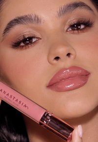 Płynna szminka w różowej tubce z różowozłotymi akcentami. Modelka ma błyszczące nude usta i neutralny makijaż oczu, podkreślający kolor produktu.