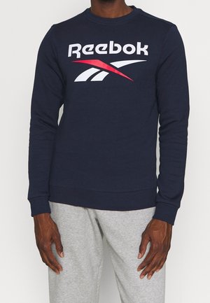 Mies pukeutuneena tummansiniseen Reebokin collegepaitaan, jossa valkoinen ja punainen logo, yhdistettynä vaaleanharmaisiin collegehousuihin, seisoo yksinkertaisen taustan edessä.